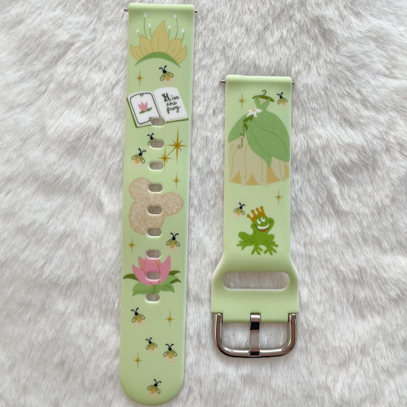 May include: Two green and white patterned watch bands with a princess and frog design. The band on the left has the text "Kiss the frog" on it. The band on the right has a silver buckle.