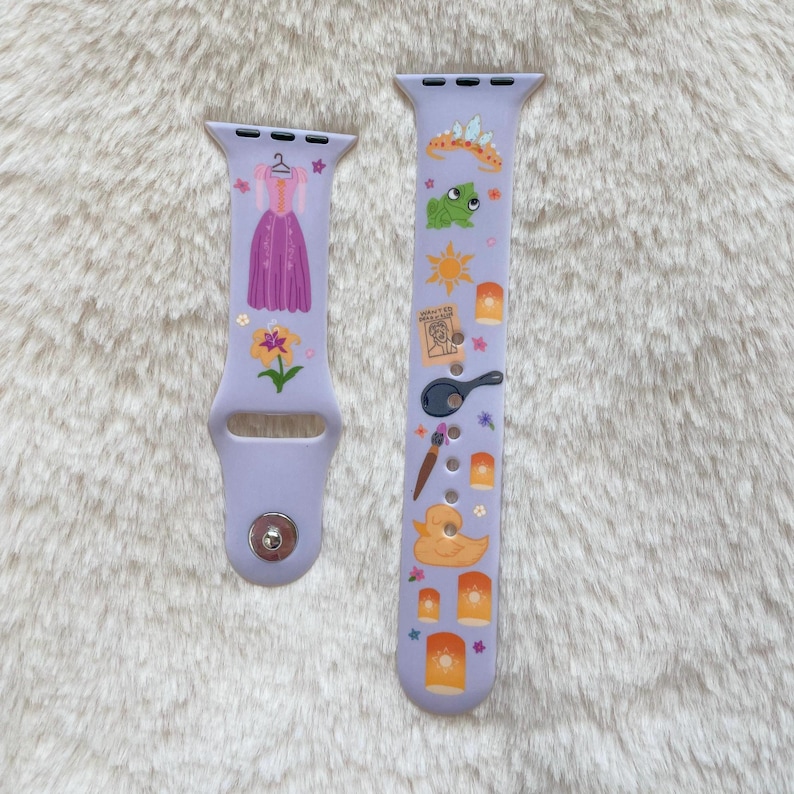 May include: Two lavender watch bands with colorful graphics. One band features a purple dress, flowers, and stars. The other band has a frog, sun, lanterns, a frying pan, and a duck. The bands are made of a flexible material.