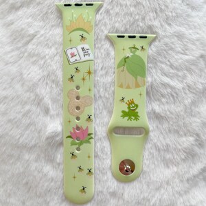 May include: A green and white Apple Watch band with a princess and frog design. The band features a green frog with a crown, a princess in a green dress, and the text "Kiss are frog".
