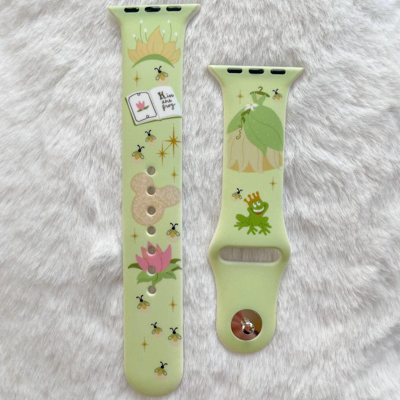 May include: A green and white Apple Watch band with a princess and frog design. The band features a green frog with a crown, a princess in a green dress, and the text "Kiss are frog".