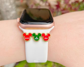 Puede incluir: Un Apple Watch blanco con una banda de oro rosa y tres dijes de Mickey Mouse. Los dijes son rojos, verdes y rojos y dicen "Ho Ho Ho", "Be Merry" y "Jolly".