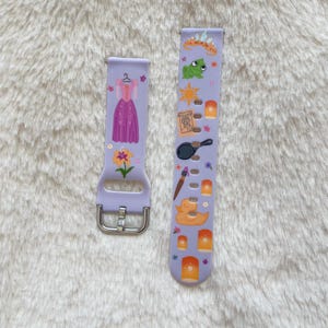 May include: Two lavender watch bands with a silver buckle. One band features a purple dress, a flower, and a hanger. The other band has a frog, sun, lanterns, a frying pan, and a paintbrush. The bands are decorated with images from a popular animated film.