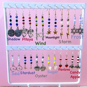 May include: A white metal earring display stand with two tiers, holding various beaded earrings. The earrings feature charms, beads, and different color combinations. Text labels include "Shadow Pitaya", "Wind", "Moonlight", "Storm", "Frost", "Sea", "Stardust", "Oyster", "Sugar", "Pavlova", and "Candy Apple".