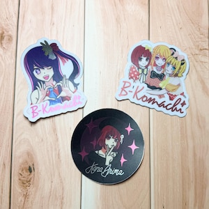 May include: Three anime-style stickers on a wooden surface. One sticker features a girl with purple hair and the text "B-Komachi". Another shows three girls and the text "B-Komachi". The third is a black circle with a girl and the text "Kana Arima".