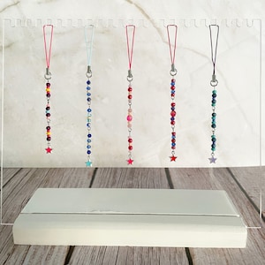 May include: Six colorful beaded phone charms with star-shaped charms at the bottom. The charms are displayed on a clear acrylic stand.