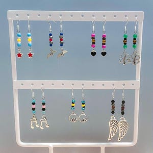 May include: A display of handmade earrings with various designs. Some earrings feature colorful beads with star charms, bat charms, and heart charms. Other earrings have musical notes, mountain scenes, and wing charms. The earrings are displayed on a white earring stand.