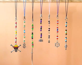 May include: A collection of beaded phone charms hanging from a wooden rod. The charms feature various designs, including a skull and crossbones, a dollar sign, a sword, a building, a small charm, a ship's wheel, and a fish.