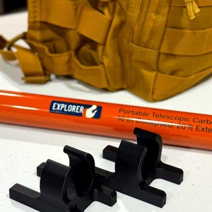May include: An orange and black portable telescopic carbon fiber pole with the word "EXPLORER" printed on it. Two black plastic brackets are in the foreground. A tan backpack is in the background.