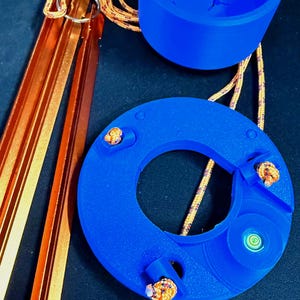 May include: A blue 3D-printed circular levelling tool with a built-in bubble level, a matching blue cylindrical cup, and three metallic orange tent stakes. The components are connected by orange and yellow speckled nylon cordage with metal rings.