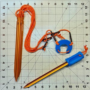 Puede incluir: Dos estacas de tienda de campaña naranjas con un anillo azul y una cuerda naranja. Las estacas son de metal y tienen un extremo puntiagudo. La cuerda está atada a las estacas y tiene un lazo en el extremo. El anillo es de plástico y tiene un agujero en el centro.