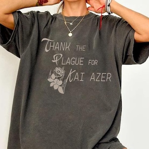 Może przedstawiać: Duży, ciemnoszary T-shirt z nadrukiem "Thank the Plague for Kai Azer" na przodzie. Tekst jest w stylizowanym foncie z grafiką róży.