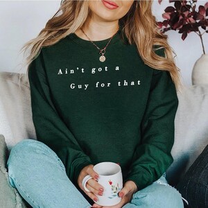 May include: A dark green sweatshirt with the text "Ain't got a Guy for that" in white. The person is holding a white mug with a floral design. The person is wearing blue jeans and a gold necklace.