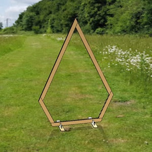 May include: A wooden geometric arch with a hexagonal shape, standing on small wooden supports. The arch is set in a grassy field with a path leading into the distance, surrounded by trees and wildflowers. The arch is a light brown color.