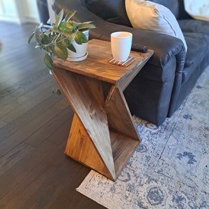Puede incluir: Una mesa auxiliar de madera con un diseño único en forma de X. La mesa tiene un acabado marrón claro y está hecha de madera maciza. Una pequeña planta en maceta y una taza blanca están sobre la mesa.