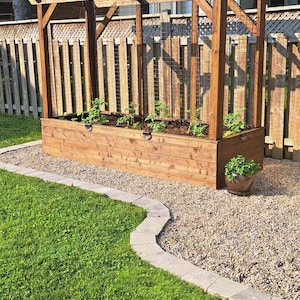 May include: A wooden garden planter with a trellis, filled with soil and young plants. The structure is stained a warm brown and sits on a bed of gravel, bordered by a stone walkway and green grass.