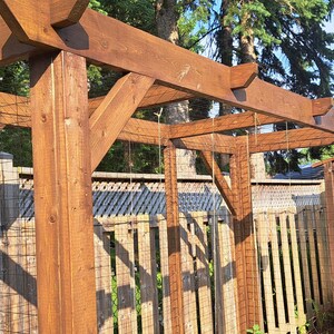 May include: A wooden pergola with a lattice roof and support beams. The structure is made of stained wood and is set in a garden. A wooden fence and a small wooden shed are in the background.