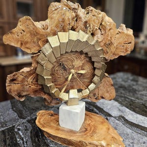 May include: A gold clock face with a geometric design is mounted on a wooden base. The base is shaped like a tree branch and has a natural wood grain. The clock is sitting on a white marble base.