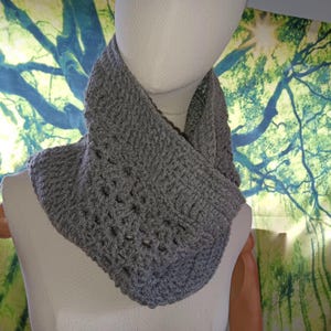 May include: A gray crocheted cowl scarf displayed on a mannequin. The scarf is made of a thick, textured yarn and has a wide, open design. The cowl is a neutral color, suitable for various outfits.