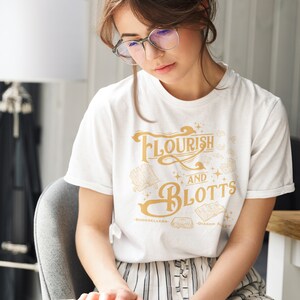 Puede incluir: Camiseta blanca con texto dorado "Flourish and Blotts - Booksellers - Diagon Alley". La persona lleva gafas y sostiene un libro abierto, sentada en una silla gris.