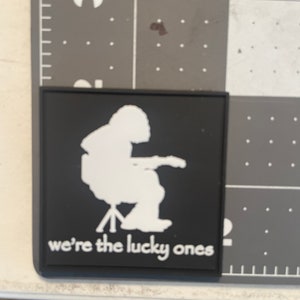 May include: Black patch with a white silhouette of a person sitting in a chair and playing a guitar. The text "we're the lucky ones" is printed below the image.