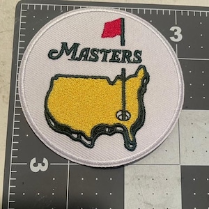 May include: White embroidered patch with the words "Masters" and a green and yellow outline of the United States. A red flag with a white pole is above the word "Masters".