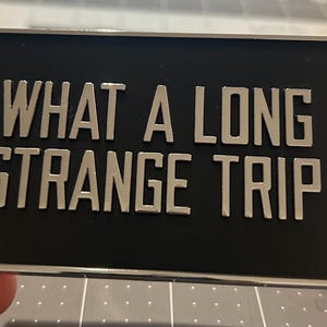 May include: A rectangular black metal pin with the phrase "WHAT A LONG STRANGE TRIP" in silver lettering. The pin is held by a hand, with a red tab visible on the side.