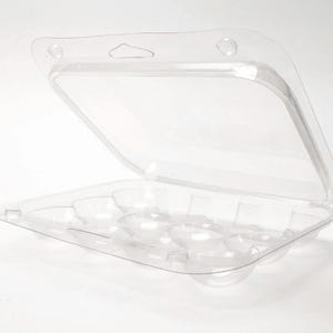 May include: A clear plastic egg carton, designed to hold six eggs. The hinged lid is open, revealing the individual egg compartments. The carton has a handle hole for easy carrying. The plastic is transparent, allowing visibility of the contents.