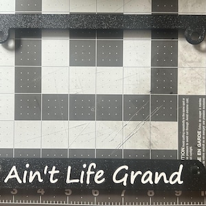 May include: A black rectangular license plate frame with a textured finish. The bottom edge features the white text "Ain't Life Grand" in a script font. The frame is positioned on a cutting mat with visible inch markings along the edges.