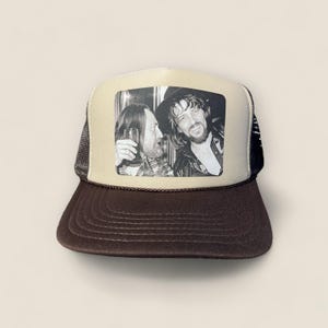 May include: A trucker hat with a brown brim and a tan front panel featuring a black and white photo of two men. The hat has a black mesh back and a brown rope detail along the brim.
