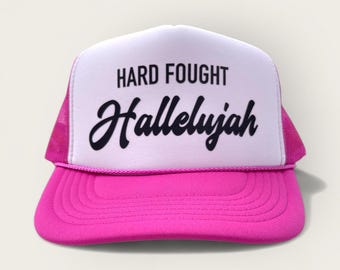 May include: A trucker hat with a white front panel and a bright pink brim and mesh back. The hat features the text "HARD FOUGHT Hallelujah" in black lettering.