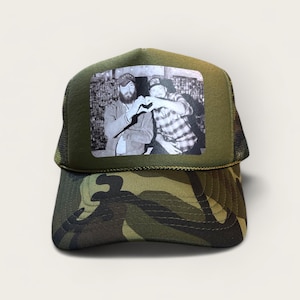 May include: A camouflage trucker hat with a black and white photo of two men making a heart shape with their hands. The photo is on the front of the hat.