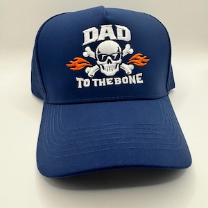 May include: A navy blue baseball cap featuring an embroidered design of a white skull wearing sunglasses, crossed bones, and orange flames. The text DAD is embroidered in white above the skull, and TO THE BONE is embroidered in white below the skull.