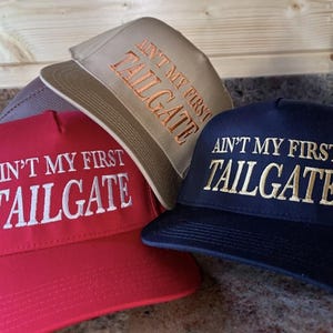 May include: Three baseball caps in red, tan, and navy blue. Each cap has the embroidered text "AIN'T MY FIRST TAILGATE" in white or gold thread. The caps are stacked, with the red cap in the foreground.