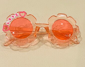 May include: Pink plastic sunglasses with a flower-shaped frame and orange lenses. The sunglasses have the word "LOVE" spelled out in pink letters on the left lens.