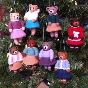May include: A collection of handmade felt teddy bear ornaments. Each bear is dressed in a different colored sweater and skirt, with white accents. The ornaments are hanging on a Christmas tree, creating a festive holiday display.