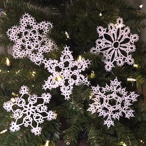 May include: Five white crocheted snowflakes hanging on a green Christmas tree with white lights.