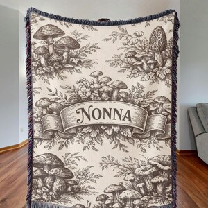 May include: A cream-colored woven throw blanket featuring a dark brown botanical illustration of various mushrooms and foliage. A central banner displays the text NONNA in a decorative font. The blanket has a dark fringe along all four edges.