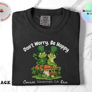 May include: Black crew neck t-shirt with the text "Don't Worry, Be Hoppy" and a cartoon frog design. The frogs are wearing green hats and sitting on a mushroom. The shirt also has the text "Connor Savannah, GA Erin".