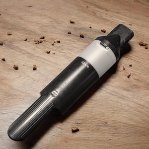 May include: A black and white handheld vacuum cleaner with the word "VYE" printed on the side. The vacuum cleaner is lying on a wooden surface.