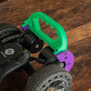 Könnte beinhalten: Eine elektrische Skateboard-Radbaugruppe mit einem grünen Griff und violetten Montagehalterungen. Das schwarze Rad hat einen profilierten Reifen. Die Baugruppe befindet sich auf einer Holzoberfläche.