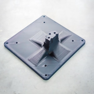 May include: A dark gray, 3D-printed base with a diamond shape. The base has a central support structure and several holes for mounting. The surface has a textured, layered appearance, suggesting a 3D printing process.