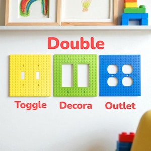 K&ouml;nnte beinhalten: Drei farbenfrohe Wandplatten im Lego-Stil: gelber Schalter, gr&uuml;ner Decora und blaue Steckdose. Das Wort "Double" steht &uuml;ber den Platten, mit den W&ouml;rtern "Toggle", "Decora" und "Outlet" darunter. Kunstwerke und Legosteine befinden sich auf einem Regal dar&uuml;ber.
