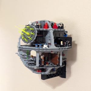May include: A gray and black Lego Death Star model, mounted on a wall. The model is partially cut away, revealing interior details and miniature figures. Green laser beams emanate from the superlaser. The model is a detailed replica of the iconic space station.