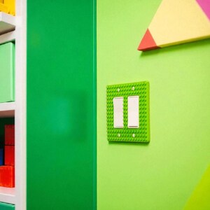 K&ouml;nnte beinhalten: Eine hellgr&uuml;ne Lichtschalterabdeckung mit zwei wei&szlig;en Schaltern, die wie Spielzeugbausteine gestaltet ist, ist an einer hellgr&uuml;nen Wand angebracht. Eine gr&uuml;ne T&uuml;r und eine farbenfrohe geometrische Form sind ebenfalls zu sehen.