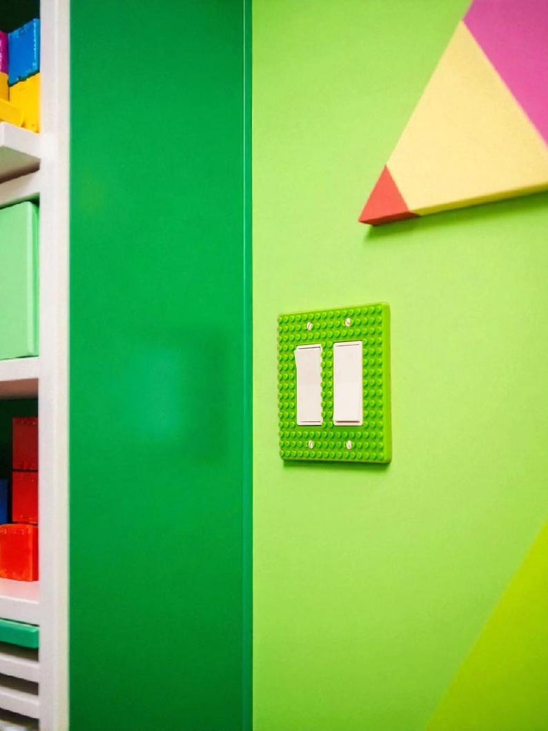 K&ouml;nnte beinhalten: Eine hellgr&uuml;ne Lichtschalterabdeckung mit zwei wei&szlig;en Schaltern, die wie Spielzeugbausteine gestaltet ist, ist an einer hellgr&uuml;nen Wand angebracht. Eine gr&uuml;ne T&uuml;r und eine farbenfrohe geometrische Form sind ebenfalls zu sehen.