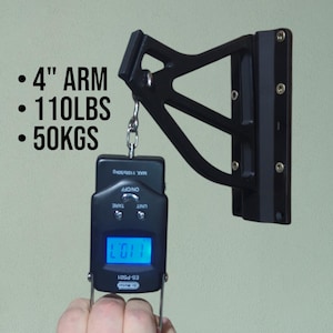 May include: A black wall-mounted bracket with a 4-inch arm, supporting a digital scale. The scale displays a weight of 110.7, with a maximum capacity of 110 lbs or 50 kgs. The scale has an on/off button and a tare function.