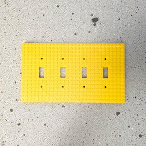 K&ouml;nnte beinhalten: Eine leuchtend gelbe, rechteckige Lichtschalterabdeckung mit vier Schalter&ouml;ffnungen. Die Abdeckung besteht aus einem strukturierten Material mit kleinen kreisf&ouml;rmigen Noppen, die an ein Bausteindesign erinnern. Der Hintergrund ist eine gesprenkelte graue Oberfl&auml;che.