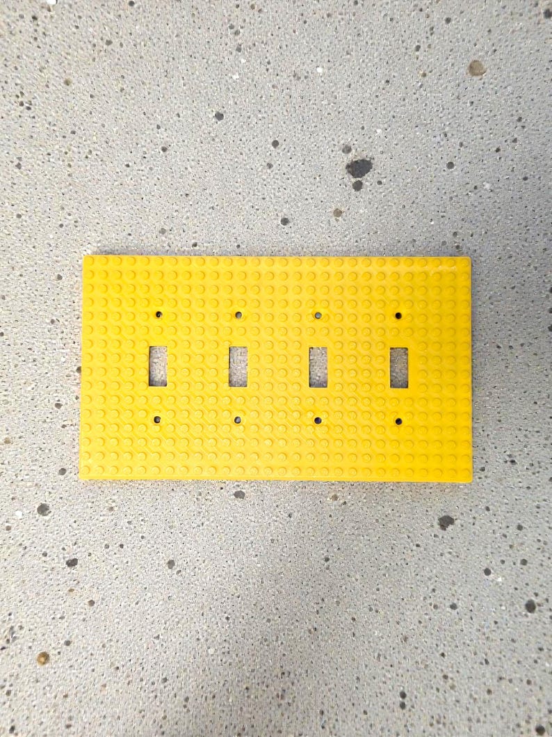 K&ouml;nnte beinhalten: Eine leuchtend gelbe, rechteckige Lichtschalterabdeckung mit vier Schalter&ouml;ffnungen. Die Abdeckung besteht aus einem strukturierten Material mit kleinen kreisf&ouml;rmigen Noppen, die an ein Bausteindesign erinnern. Der Hintergrund ist eine gesprenkelte graue Oberfl&auml;che.