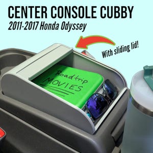 May include: A gray center console cubby with a sliding lid. The cubby is open and contains a green book with the words "Roadtrip Movies" written on it and a pair of sunglasses. The text "2011-2017 Honda Odyssey" is written above the cubby. The text "With sliding lid!" is written in the upper right corner of the image.