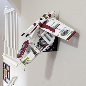 May include: A white, red, and gray Lego spaceship model mounted on a black bracket against a white wall. The model features wings, engines, and a cockpit. The model is a detailed replica of a starship.